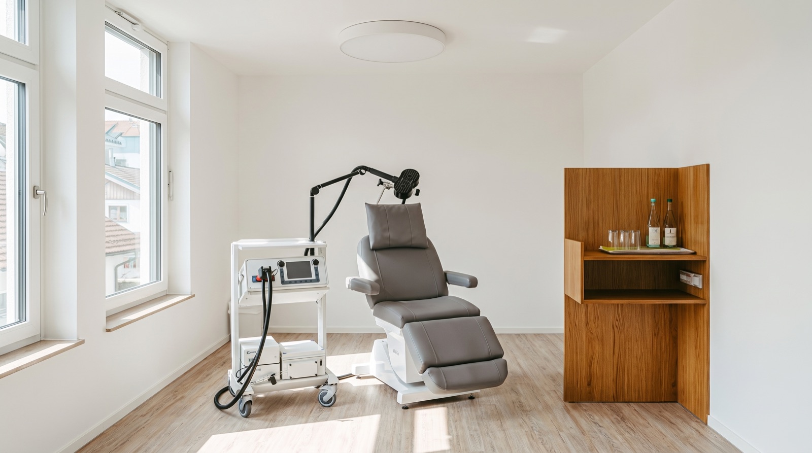 Treatment room with a grey reclining chair, a white rTMS cart with articulated arm and figure-8 coil, an oak cabinet and tall windows in morning light