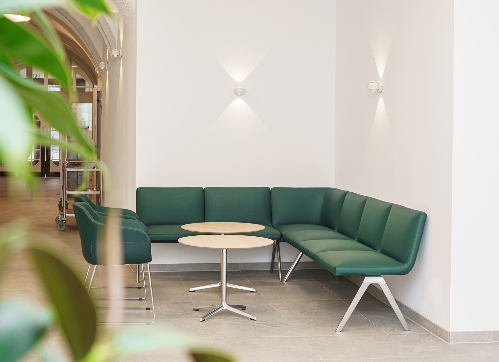 The entrance area of Clinicum St. Georg: green bench, round oak tables, plants and warm light.