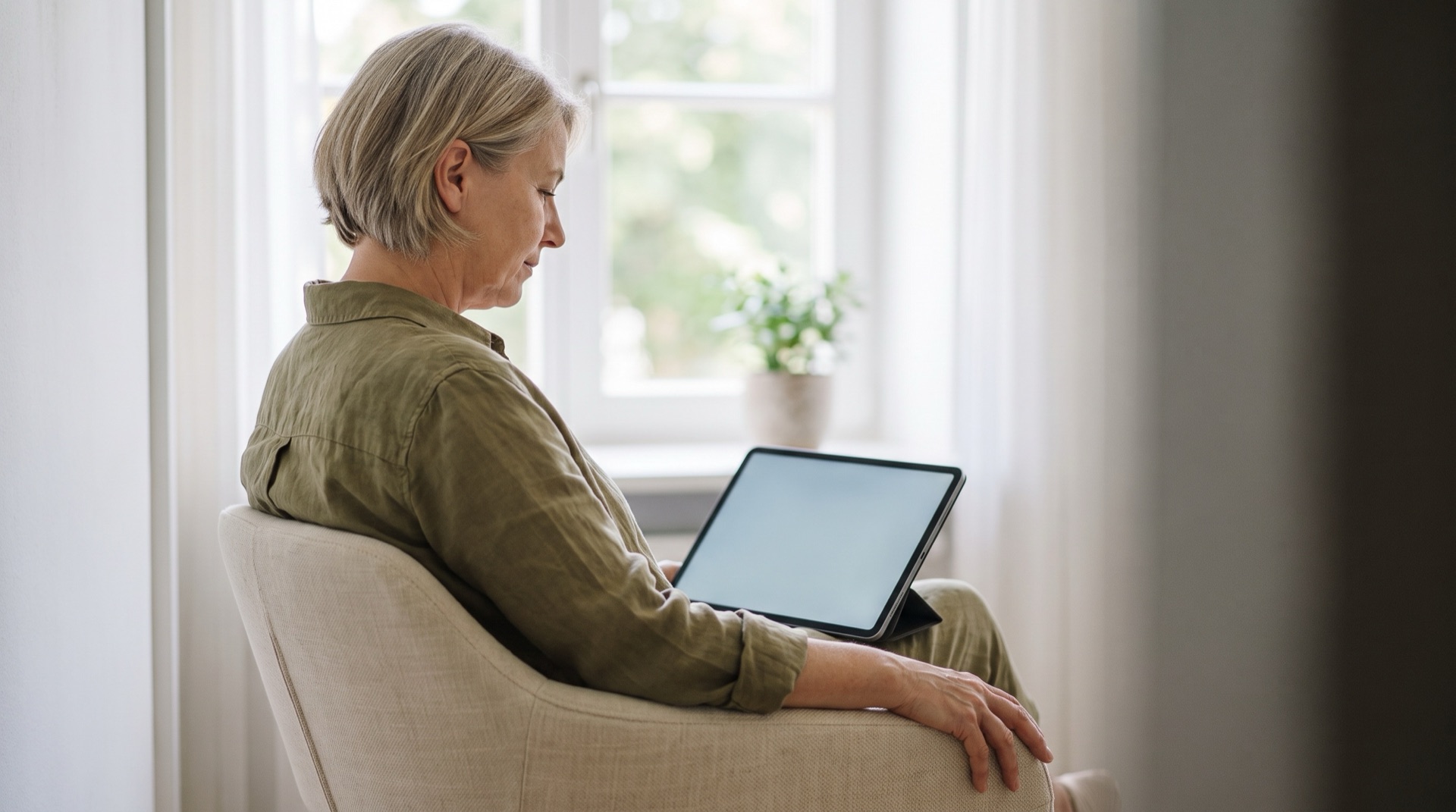 A woman in her mid-fifties sits in an armchair by the window, a tablet on her lap, reading calmly.