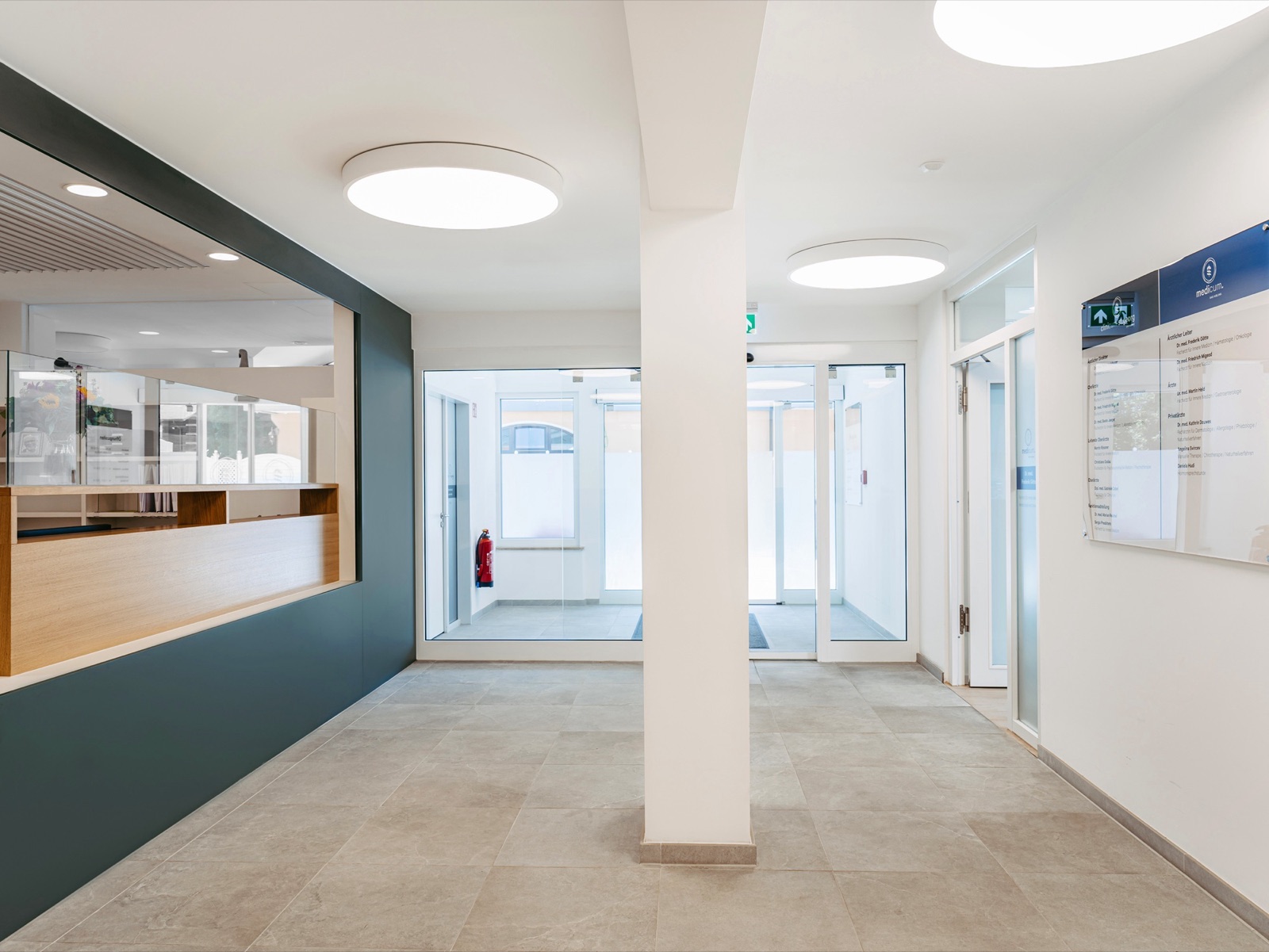 The reception of Clinicum St. Georg: oak counter, dark green accent wall, round ceiling lights, glass doors.