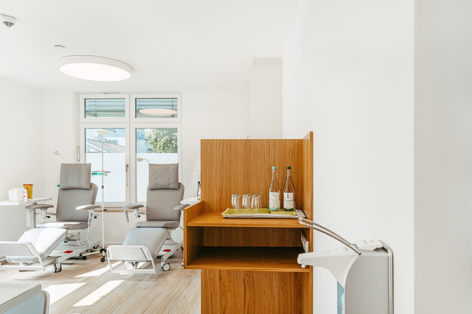 Treatment room with white walls, light wooden floor, tall windows and grey treatment chairs.
